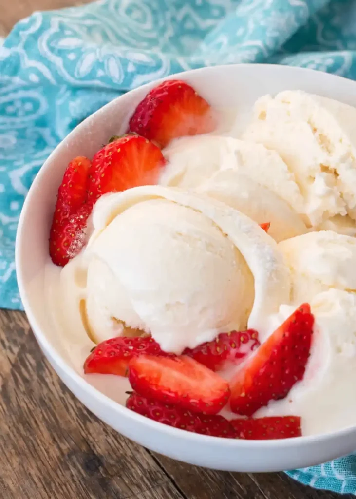 Bowl of vanilla ice cream with fresh sliced strawberries on a wooden table with a blue patterned cloth.