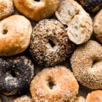 Assorted homemade bagels with different toppings including sesame, poppy seed, everything seasoning, and sugar crust, some sliced to show a soft interior