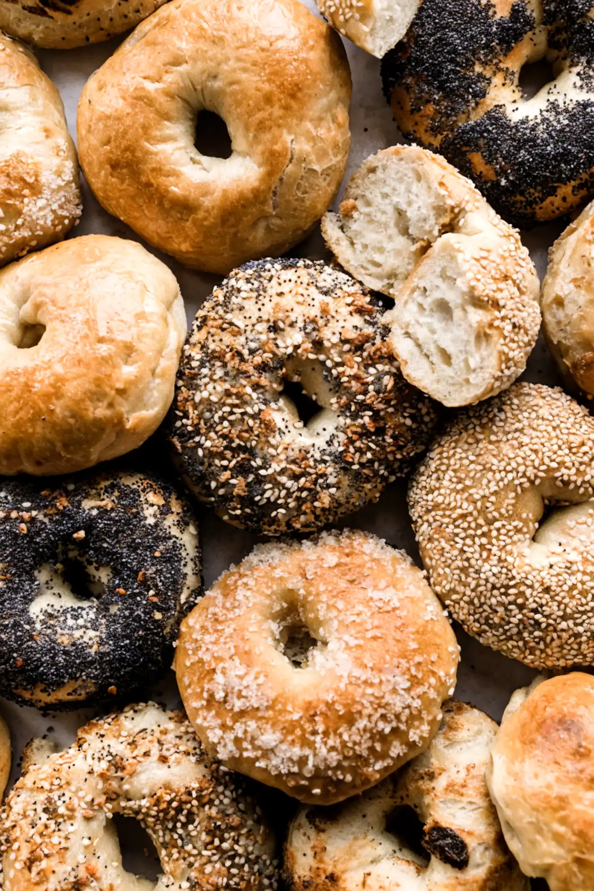 Assorted homemade bagels with different toppings including sesame, poppy seed, everything seasoning, and sugar crust, some sliced to show a soft interior