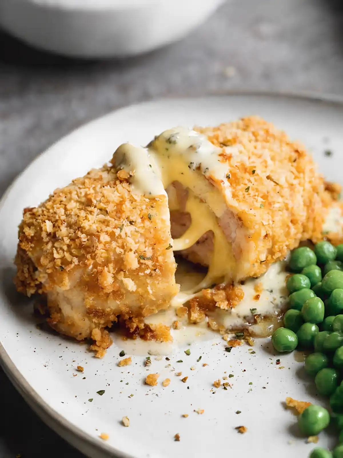Baked chicken cordon bleu with crispy golden coating, sliced open to reveal melted cheese and ham, served with green peas on a white plate.