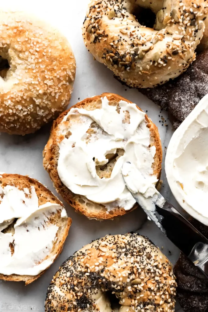 Toasted bagel spread with cream cheese surrounded by assorted homemade bagels with sesame, everything seasoning, and sugar toppings