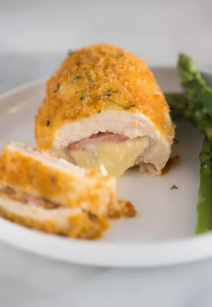 Crispy baked chicken cordon bleu sliced open with melted cheese and ham filling, served with asparagus on a white plate.