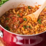 Easy goulash in a red pot with macaroni, ground beef, and tomato sauce, stirred with a wooden spoon