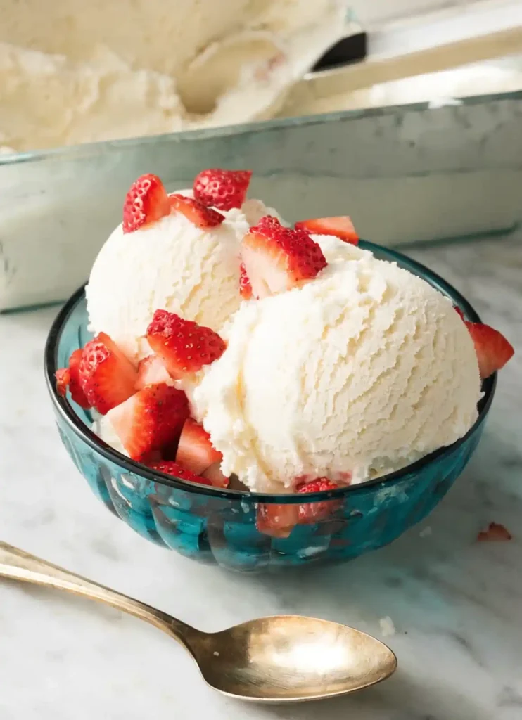Teal bowl of homemade vanilla ice cream topped with fresh strawberries beside a gold spoon on a marble surface.