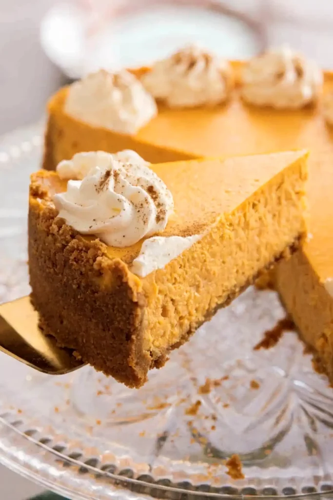Slice of pumpkin cheesecake with graham cracker crust and whipped cream on a cake stand