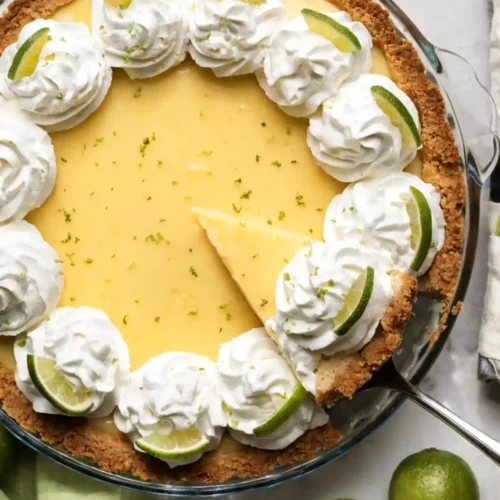 Key lime pie with whipped cream topping, lime slices, and graham cracker crust with a slice being served