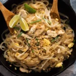 Chicken pad thai in a skillet with rice noodles, bean sprouts, eggs, peanuts, and lime wedges