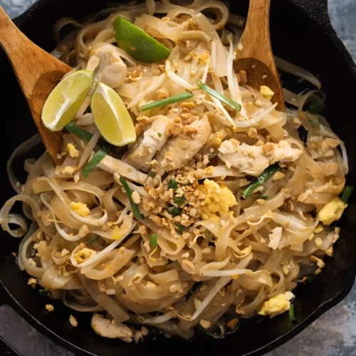 Chicken pad thai in a skillet with rice noodles, bean sprouts, eggs, peanuts, and lime wedges