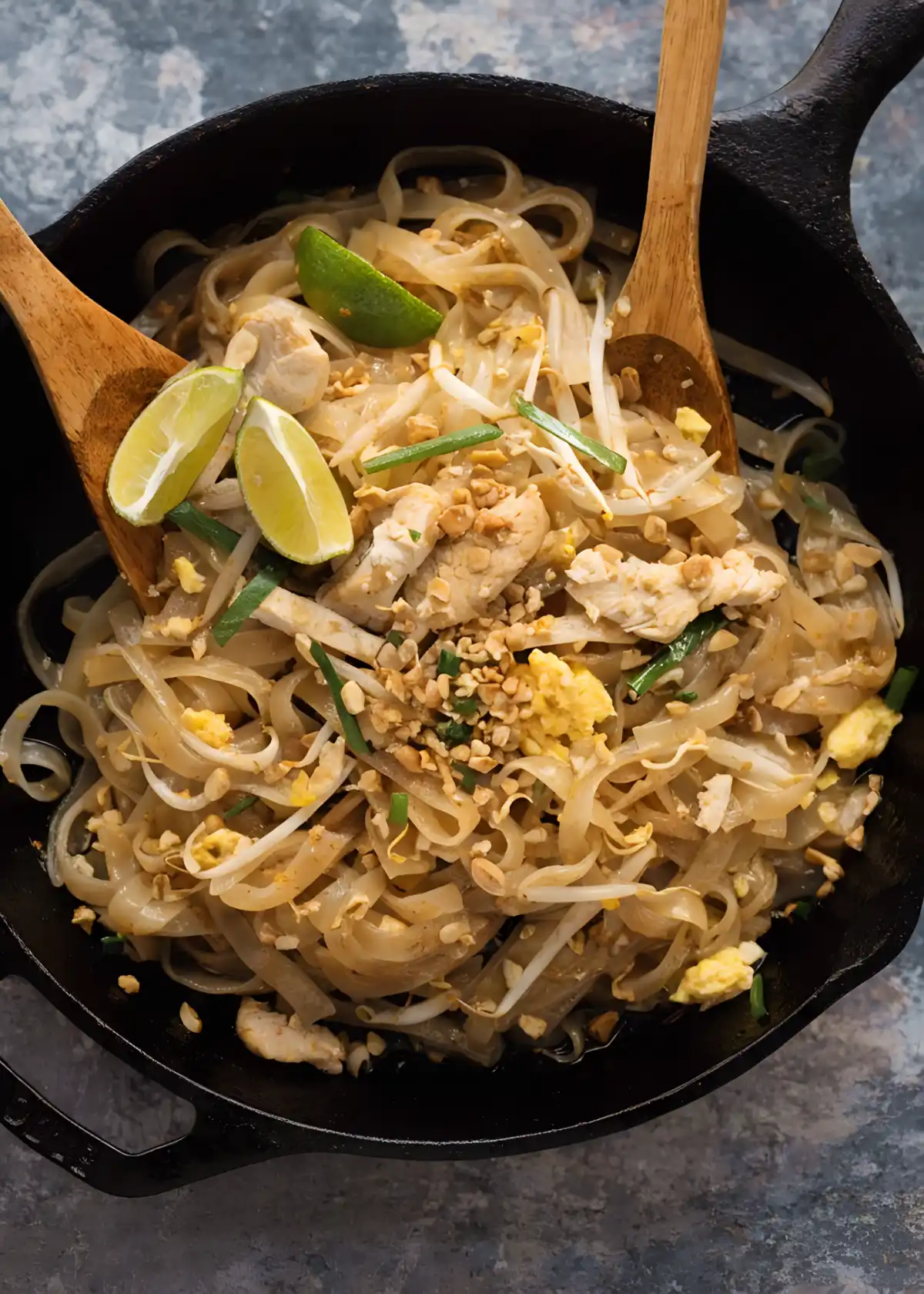 Chicken pad thai in a skillet with rice noodles, bean sprouts, eggs, peanuts, and lime wedges