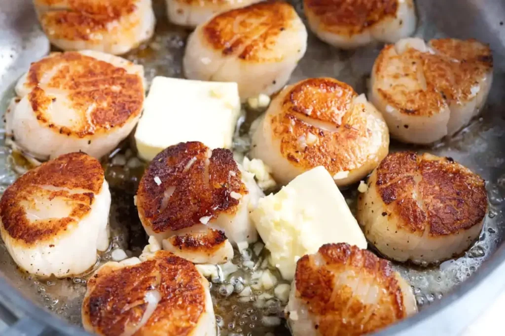 Pan seared scallops in a hot skillet with golden crust, melting butter cubes, and minced garlic sizzling around