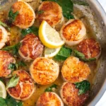 Pan seared scallops with golden crust in garlic butter sauce, topped with fresh basil leaves and lemon slices in a skillet