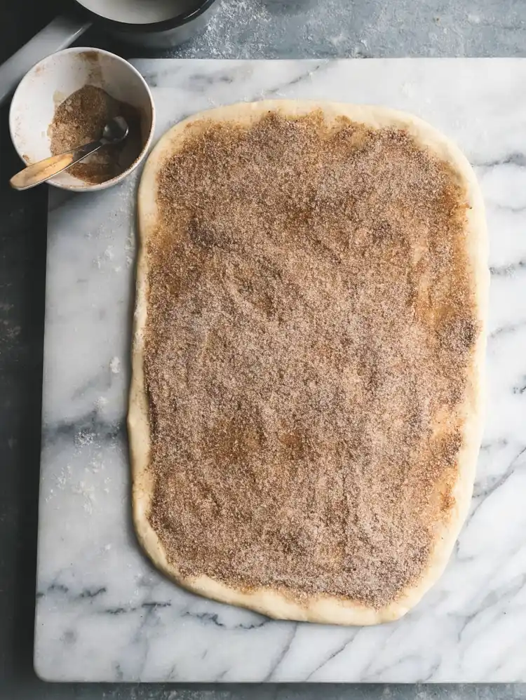 Sourdough cinnamon roll dough rolled flat and covered with cinnamon sugar filling on a marble surface