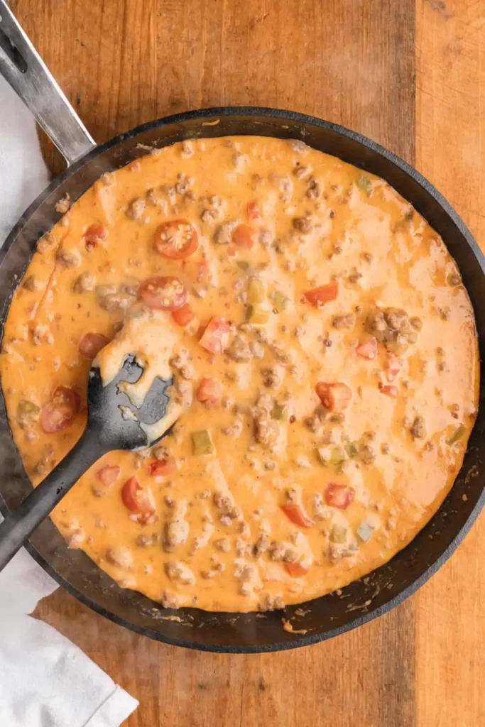 Creamy velveeta rotel dip cooking in a skillet with ground beef, tomatoes, and green chilies on a wooden table.