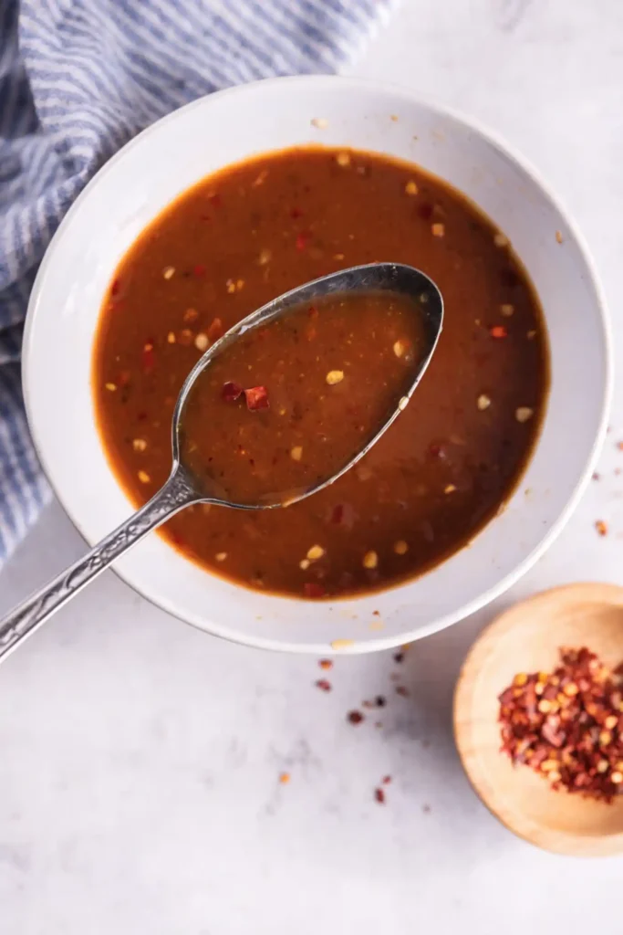 Spoon lifting homemade stir fry sauce from a white bowl with red pepper flakes on a light countertop next to a striped towel.