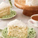 Slice of moist coconut cake on a green speckled plate with tea, flowers, and a full coconut layer cake on a wooden stand in the background.