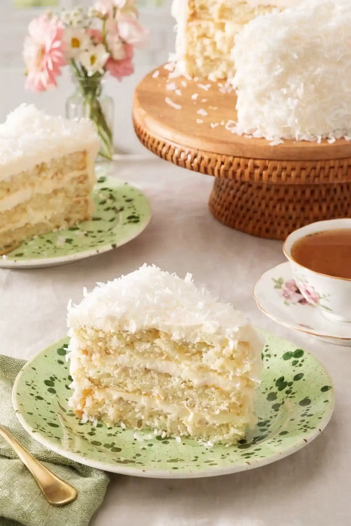 Slice of moist coconut cake on a green speckled plate with tea, flowers, and a full coconut layer cake on a wooden stand in the background.