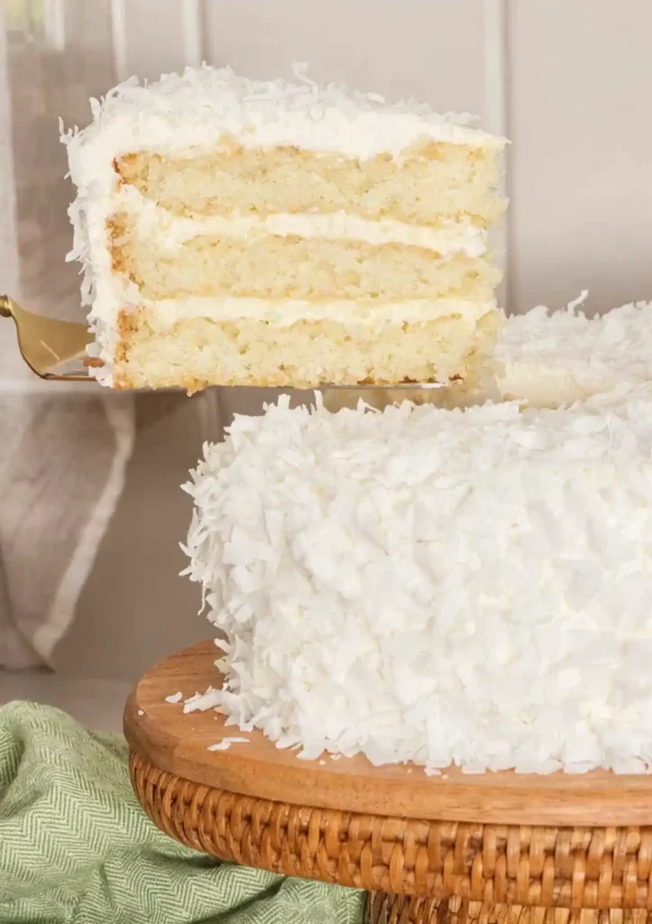 Slice of coconut layer cake being lifted from a white frosted cake covered in shredded coconut on a wooden cake stand.