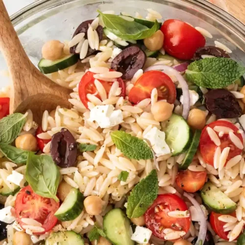 Mediterranean orzo salad in a glass bowl with cucumbers, cherry tomatoes, chickpeas, feta cheese, Kalamata olives, fresh basil, mint, and a wooden spoon.
