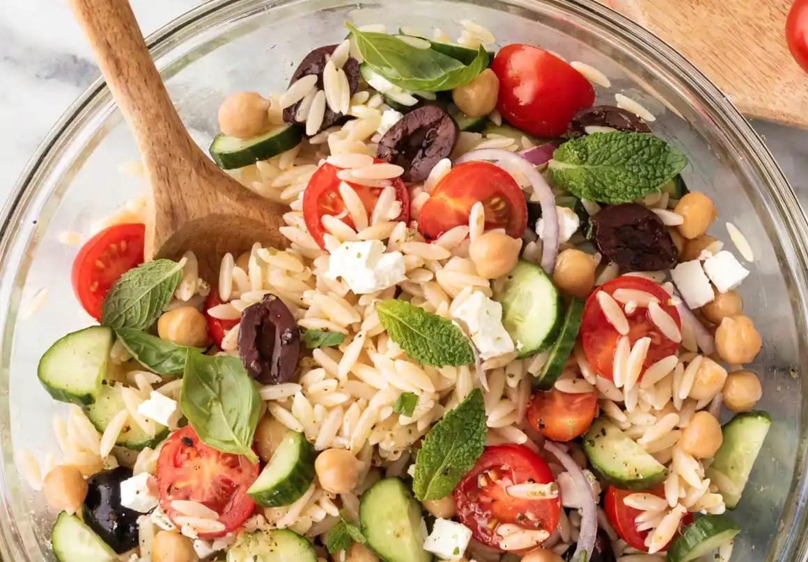 Mediterranean orzo salad in a glass bowl with cucumbers, cherry tomatoes, chickpeas, feta cheese, Kalamata olives, fresh basil, mint, and a wooden spoon.