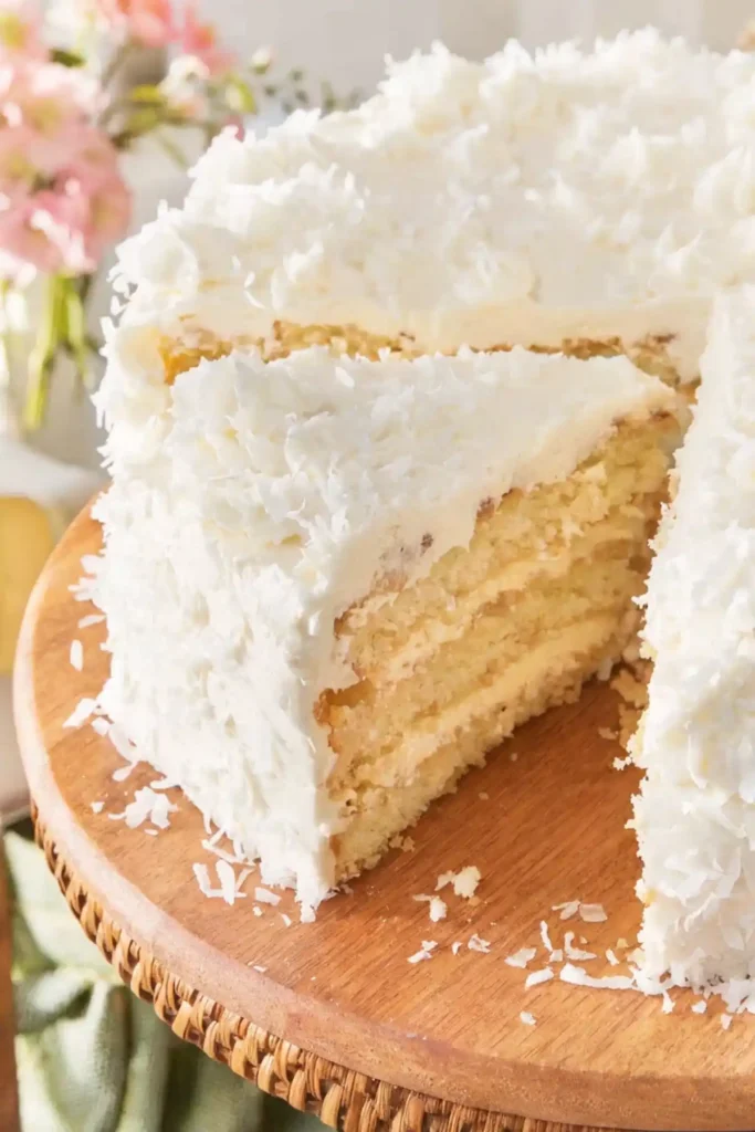Close-up of a moist coconut cake with a slice removed, showing soft cake layers and cream cheese frosting covered in shredded coconut.