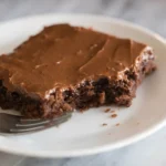 Slice of Texas sheet cake on a white plate with chocolate frosting and a bite taken out