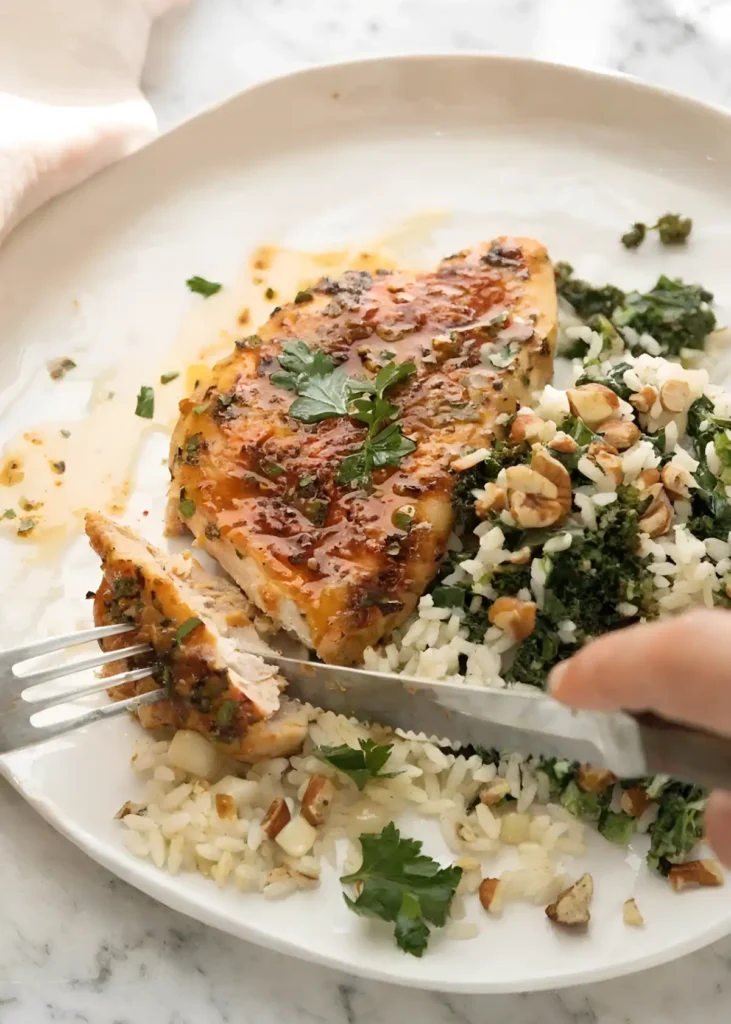 Baked chicken breasts served with rice, kale, and chopped nuts on a white plate while being sliced with a fork and knife.