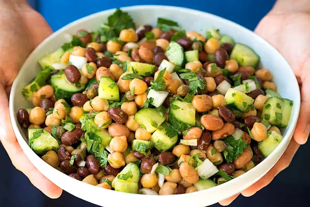 Person holding a white bowl filled with fresh bean salad made with chickpeas, black beans, pinto beans, cucumber, onions, capers, and parsley.