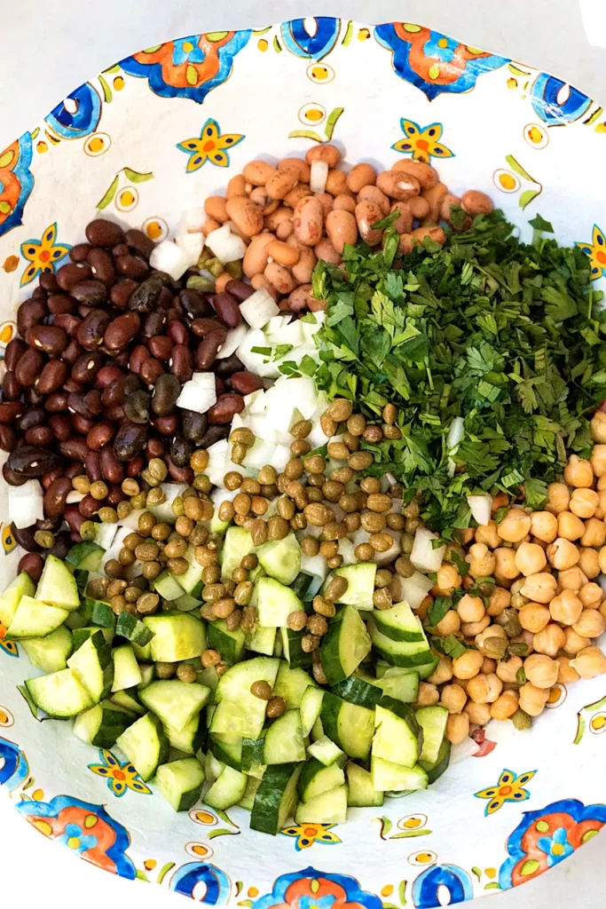 Overhead view of bean salad ingredients in a colorful bowl with black beans, chickpeas, pinto beans, cucumber, onions, capers, and fresh parsley.
