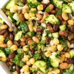 Close-up of homemade bean salad with chickpeas, black beans, pinto beans, cucumber, onions, capers, and fresh parsley in a white bowl.