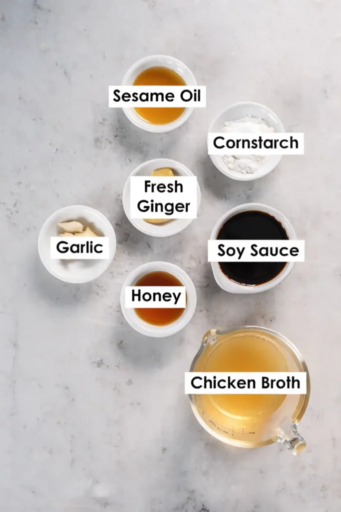 Stir fry sauce ingredients in small white bowls labeled sesame oil, cornstarch, fresh ginger, garlic, soy sauce, honey, and chicken broth on a light countertop.