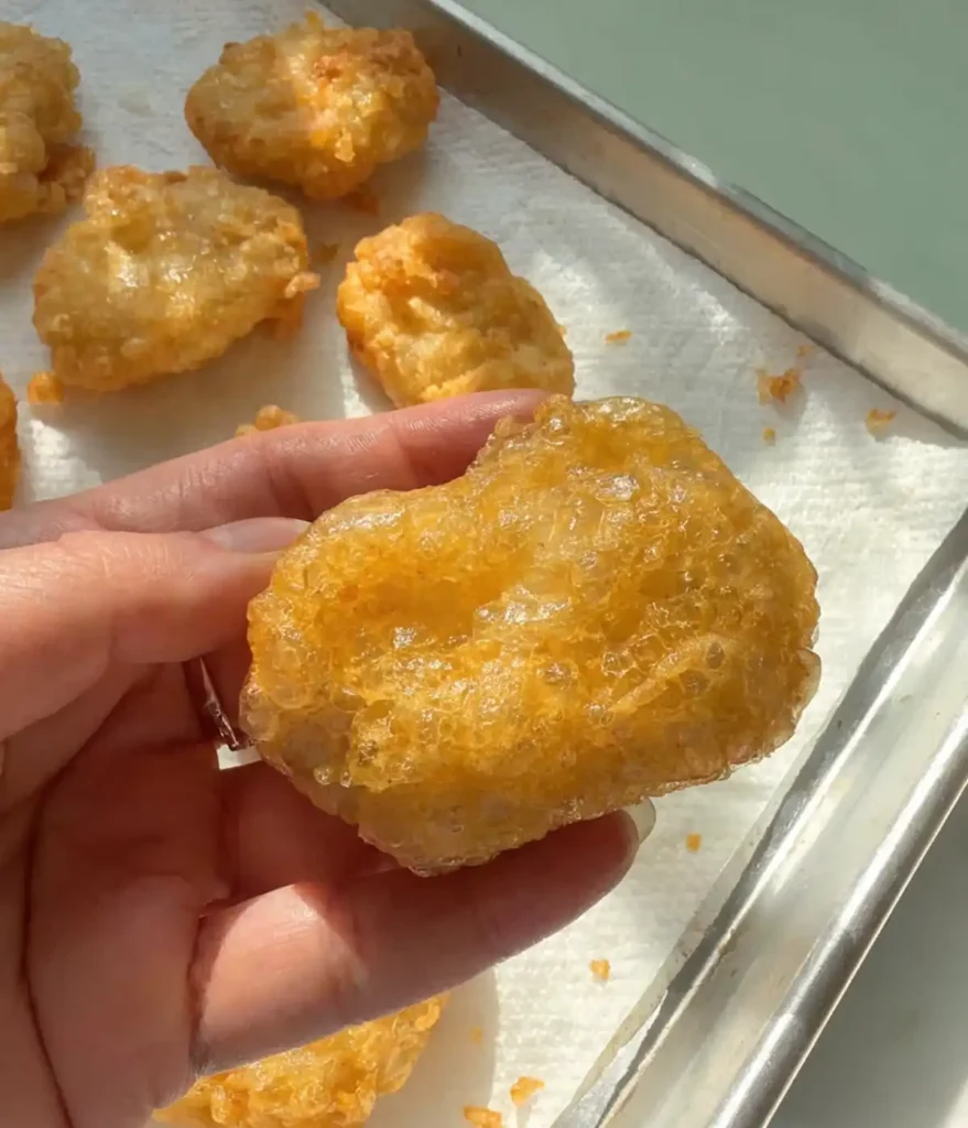 Hand holding a crispy homemade chicken nugget over a tray of freshly fried golden chicken nuggets.