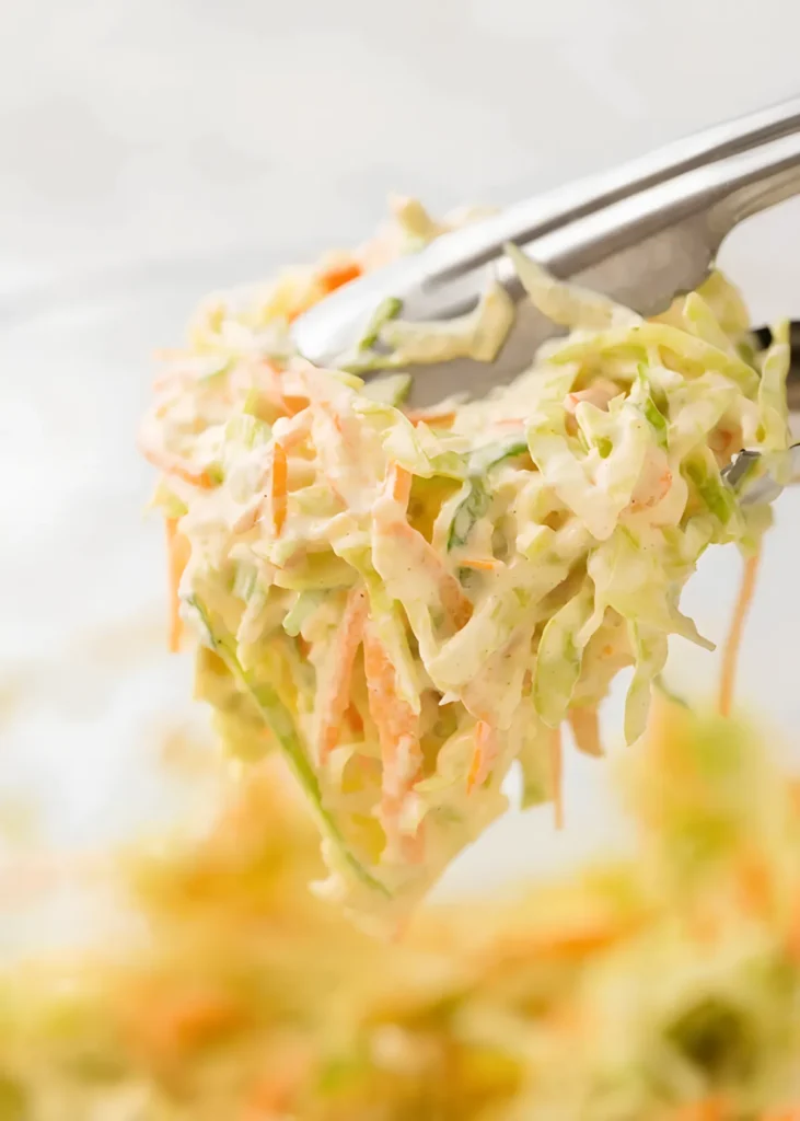 Creamy coleslaw recipe being lifted with metal tongs, showing shredded cabbage and carrots coated in a rich homemade dressing.