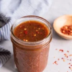 Homemade stir fry sauce in a glass mason jar with red pepper flakes on a light countertop beside a striped kitchen towel and small wooden bowl.