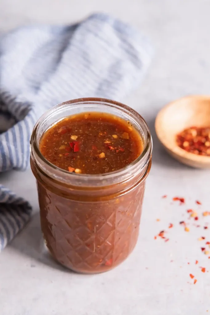 Homemade stir fry sauce in a glass mason jar with red pepper flakes on a light countertop beside a striped kitchen towel and small wooden bowl.