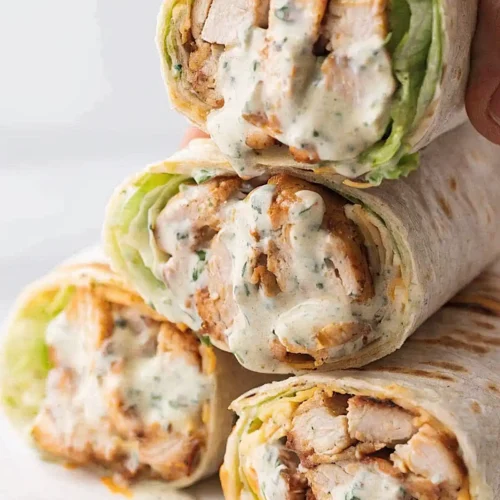 Stacked grilled chicken wraps filled with lettuce, shredded cheese, and creamy ranch sauce on a white plate.