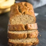 Healthy banana bread loaf with sliced pieces stacked in front on a dark countertop with ripe bananas in the background.