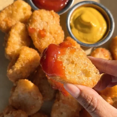Hand holding a crispy homemade chicken nugget dipped in ketchup with mustard, ketchup bowls, and golden fried nuggets in the background.