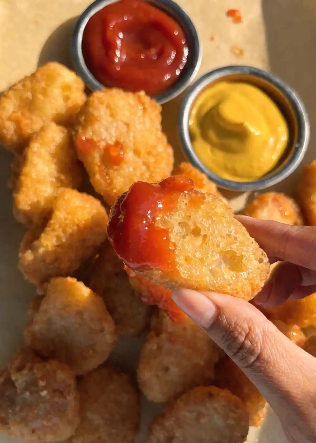 Hand holding a crispy homemade chicken nugget dipped in ketchup with mustard, ketchup bowls, and golden fried nuggets in the background.
