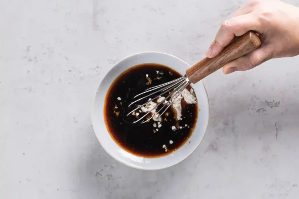 Hand whisking homemade stir fry sauce in a white bowl with soy sauce, cornstarch, and garlic on a light countertop.