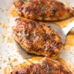 Juicy baked chicken breasts in a baking dish with caramelized seasoning glaze and fresh parsley garnish being lifted with a spatula.
