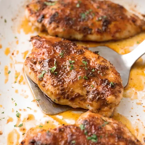 Juicy baked chicken breasts in a baking dish with caramelized seasoning glaze and fresh parsley garnish being lifted with a spatula.