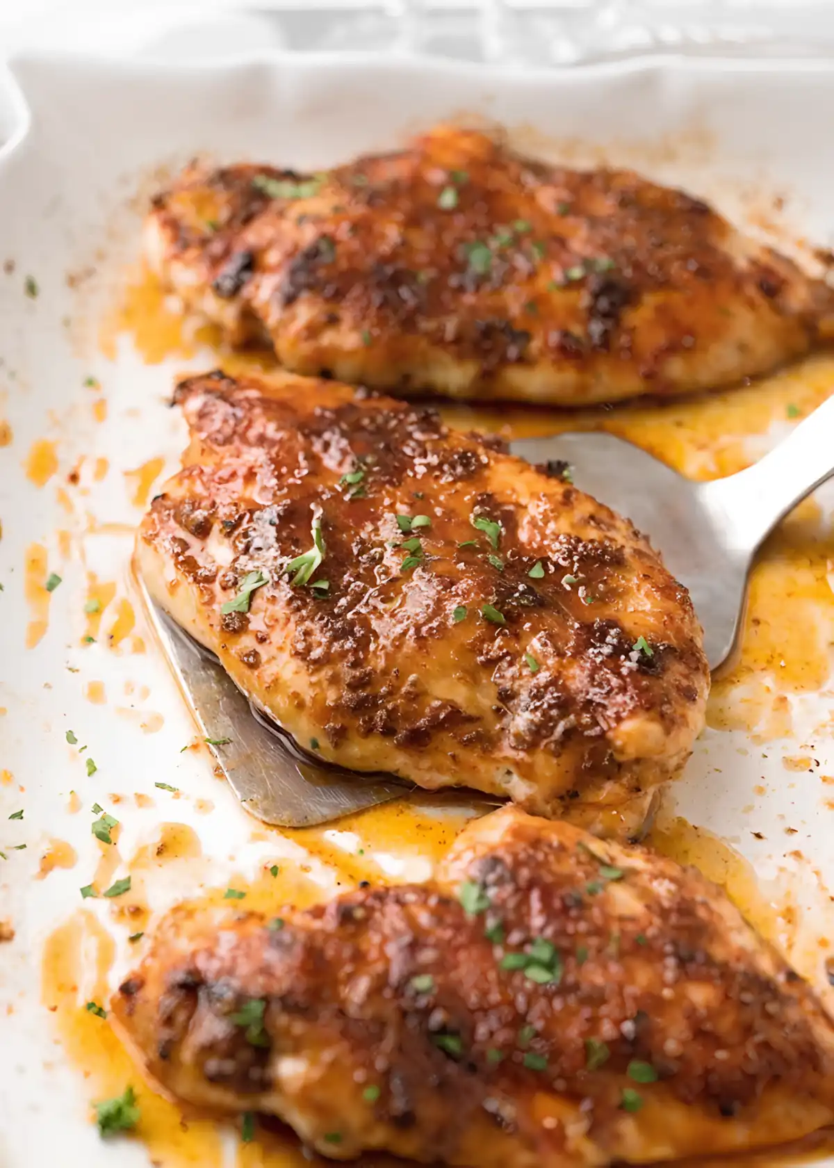 Juicy baked chicken breasts in a baking dish with caramelized seasoning glaze and fresh parsley garnish being lifted with a spatula.