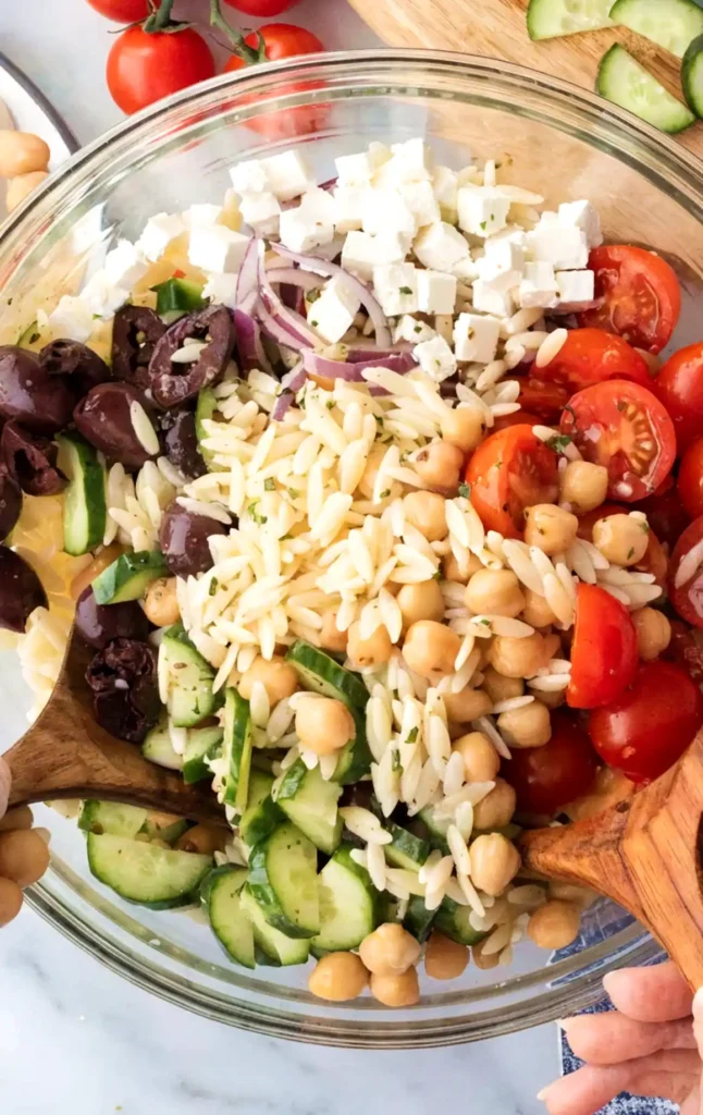 Orzo pasta salad being tossed in a glass bowl with cucumbers, cherry tomatoes, chickpeas, feta cheese, Kalamata olives, and red onions.