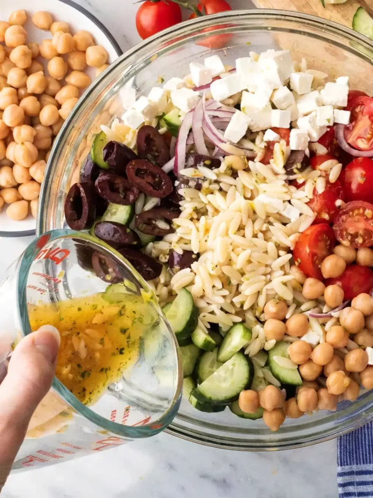 Hand pouring lemon herb dressing into a glass bowl filled with orzo salad ingredients like cucumbers, chickpeas, feta, olives, tomatoes, and red onions.