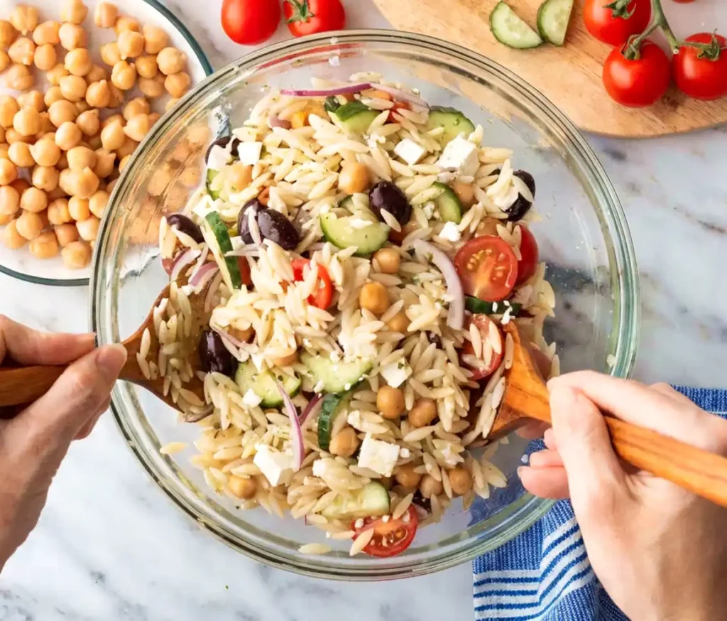 Hands tossing orzo salad in a glass bowl with cucumbers, cherry tomatoes, chickpeas, feta cheese, olives, and red onions.