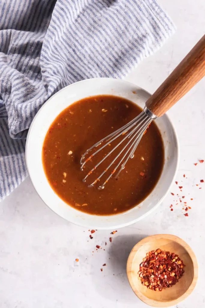 Whisk mixing homemade stir fry sauce in a white bowl with red pepper flakes on a light countertop beside a striped kitchen towel.